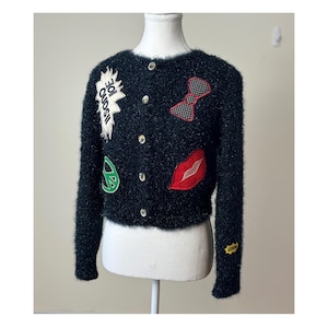 Puede incluir: Cárdigan negro y difuminado con varios parches, incluyendo una pajarita roja, labios rojos y un símbolo de la paz. El cárdigan tiene botones dorados y el texto "ISONO IOE".