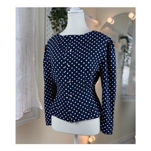 May include: A navy blue blouse with a white polka dot pattern. The long-sleeved top has a round neckline and is buttoned down the front with small, round buttons. The blouse is displayed on a mannequin.