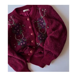 May include: A burgundy cardigan with floral embroidery and pearl detailing. The sweater features a round neck, long sleeves, and a button-down front. The floral design includes purple, green, and pink flowers.