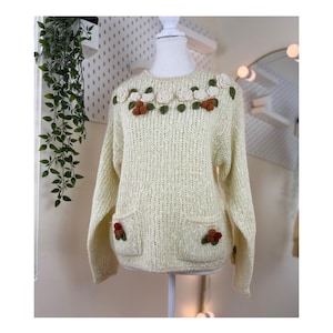 May include: Cream-coloured sweater with a round neckline and long sleeves. The sweater has two front pockets and is decorated with floral embellishments in white, green, and brown. The sweater is displayed on a mannequin.