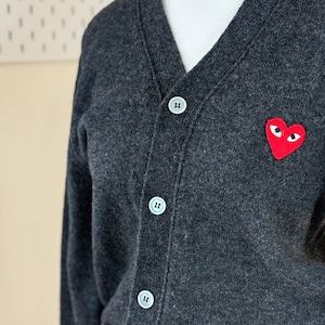 May include: Charcoal gray cardigan sweater with a V-neck and three white buttons. A red heart-shaped patch with white eyes is on the left chest. The sweater is made of a soft, textured material.