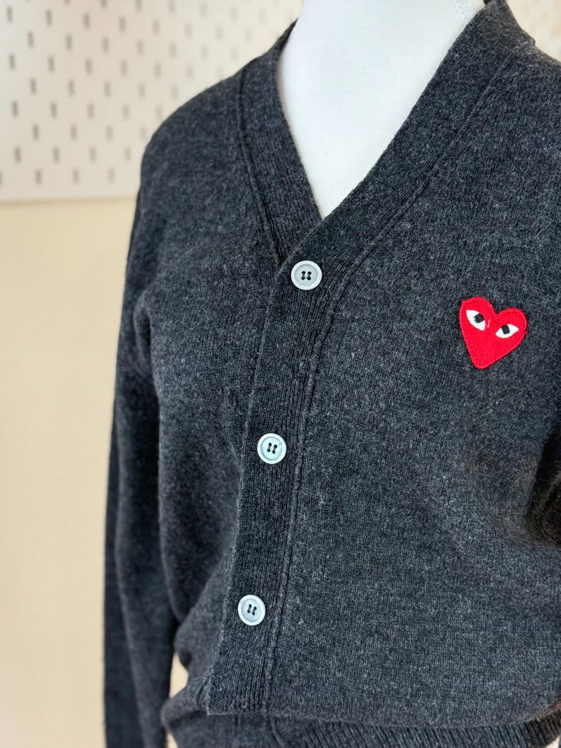 May include: Charcoal gray cardigan sweater with a V-neck and three white buttons. A red heart-shaped patch with white eyes is on the left chest. The sweater is made of a soft, textured material.