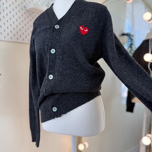 May include: Dark gray cardigan sweater with a V-neck and button closure. Features a red heart-shaped patch with eyes on the left chest. The sweater is displayed on a mannequin.