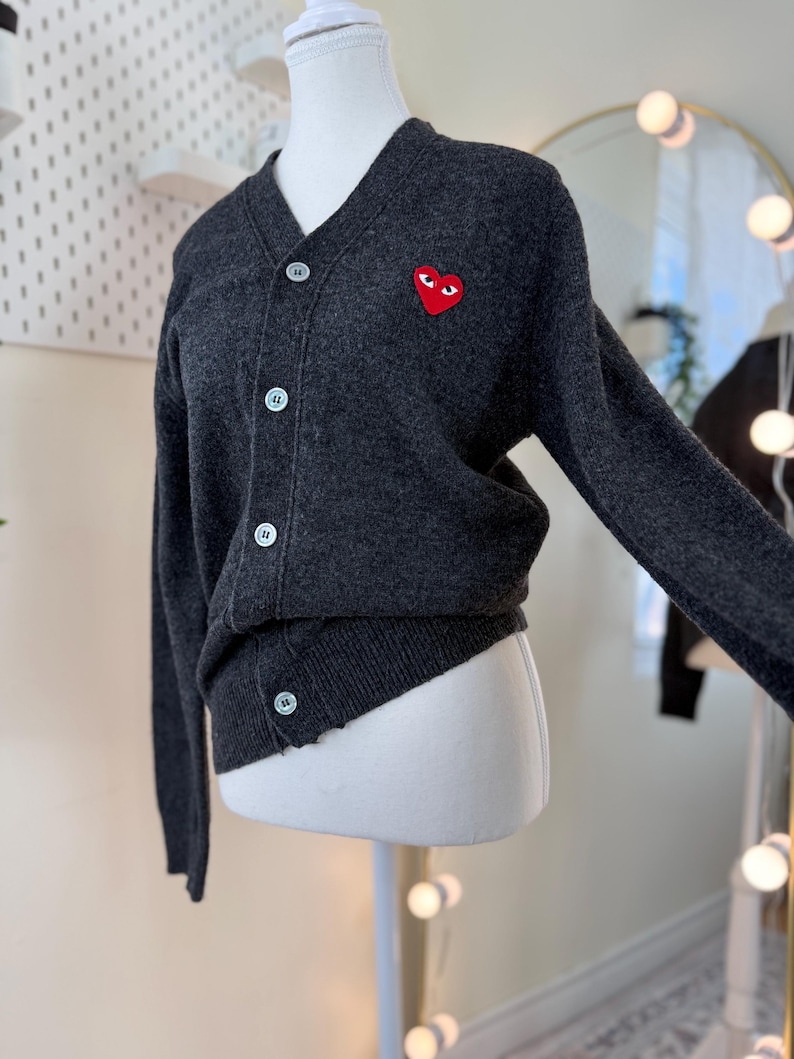 May include: Dark gray cardigan sweater with a V-neck and button closure. Features a red heart-shaped patch with eyes on the left chest. The sweater is displayed on a mannequin.