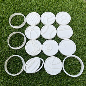 Op de afbeelding: Een verzameling witte plastic koekjessnijders in de vorm van sportballen, waaronder een golfbal, volleybal, voetbal, honkbal en basketbal. De snijders liggen op een groene ondergrond.