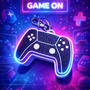Op de afbeelding: Een zwart-witte glitterende videogamecontroller-sleutelhanger met een zilveren sluiting. De controller heeft witte knoppen en een neonblauwe omtrek. De woorden "GAME ON" staan in een neonbord boven de controller.