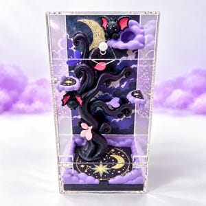 May include: A clear acrylic display box containing a dark, sculpted tree with purple and pink accents. The tree features small bat figures, a crescent moon, and star motifs. The base is decorated with purple clouds and a gold celestial design.