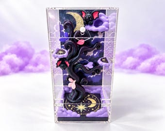 May include: A clear acrylic display box containing a dark, sculpted tree with purple and pink accents. The tree features small bat figures, a crescent moon, and star motifs. The base is decorated with purple clouds and a gold celestial design.