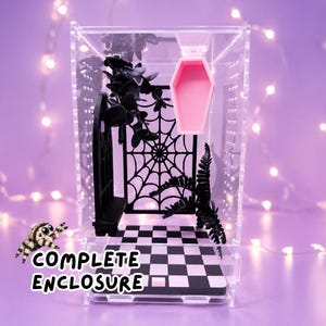 May include: A clear acrylic enclosure featuring a black and white checkered floor, a black spiderweb wall panel, a black arched window frame, a black fern, and a pink coffin-shaped hide. The text COMPLETE ENCLOSURE is printed in white.