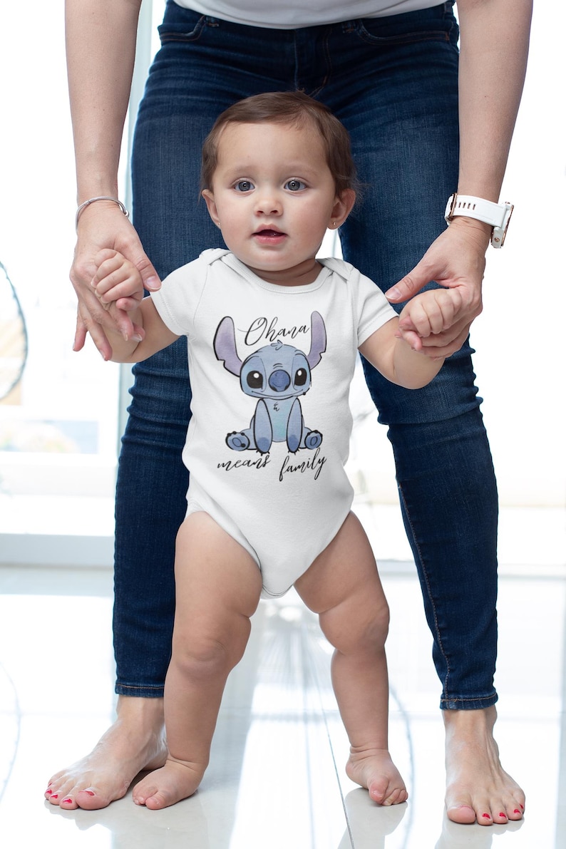 Op de afbeelding: Een witte baby romper met een blauw Stitch-personage uit de Disney-film Lilo & Stitch. De romper heeft de tekst "Ohana means family" erop gedrukt.