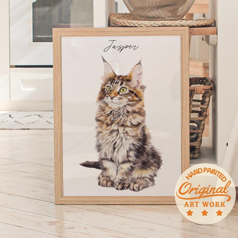 Puede incluir: Una pintura de acuarela enmarcada de un gato Maine Coon llamado Jasper. El gato tiene pelaje marr&oacute;n y canela, ojos verdes y est&aacute; sentado erguido. El marco es de color marr&oacute;n claro. La obra de arte est&aacute; etiquetada como "Hand Painted Original Art Work".