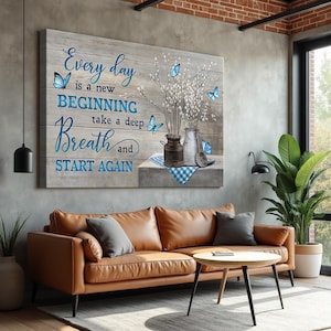 May include: A large wooden wall art piece with the text "Every day is a new beginning, take a deep breath and start again" in blue script. The art features butterflies and vases with white flowers. A brown leather sofa and a round coffee table are in front of the art.