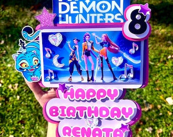 May include: A multi-layered birthday cake topper with the text "KPOP DEMON HUNTERS" and "HAPPY BIRTHDAY RENATA". The topper features a cartoon image of three characters, musical notes, and a number 8.