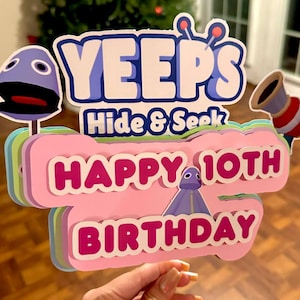 May include: A colorful layered cardboard cake topper featuring the words "YEEPS Hide & Seek Happy 10th Birthday" with a blue character and a red and white horn.