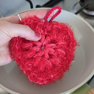 Pode incluir: Um pano de cozinha de crochê vermelho brilhante em forma de coração com um acabamento brilhante. O pano de cozinha está sendo segurado sobre uma panela de cerâmica branca.