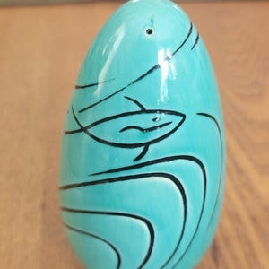 May include: A smooth, egg-shaped object in a vibrant turquoise color featuring black line art of a fish swimming among stylized waves. The surface has a glossy finish with a small hole near the top, and the overall design is simple and clean.