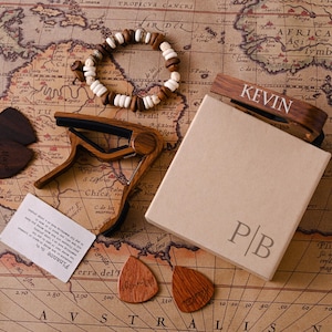 Op de afbeelding: Een assortiment houten gitaaraccessoires, waaronder plectrums, een capo en een armband, gerangschikt op een vintage kaart. Een doos met de initialen "P|B" en een capo met de inscriptie "KEVIN" zijn ook aanwezig. De items zijn bruin en beige.