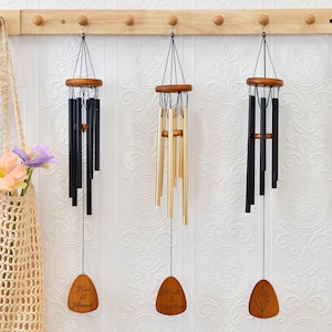 May include: Three wind chimes with wooden accents and varying tones of black and gold hang from a wooden rack. Each chime has a wooden pendant with engraved text. A beige tote bag with flowers is on the left.