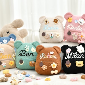 Könnte beinhalten: Eine Auswahl an teddybärförmigen Rucksäcken in verschiedenen Farben, darunter Beige, Rosa, Türkis, Braun und Schwarz. Jeder Rucksack hat einen aufgestickten Namen auf der Vorderseite sowie florale und Bären-Dekorationen. Ein Plüschbär-Spielzeug ist im Hintergrund zu sehen.