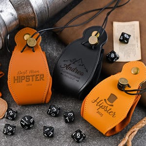 Puede incluir: Tres bolsas de dados de cuero en naranja y negro, cada una con un diseño y texto diferentes. La bolsa naranja tiene impreso "Best Man HIPSTER 1934". La bolsa negra tiene un gráfico de montaña y "Andrea Groomsman 2025.02.17". La otra bolsa naranja tiene "Hipster 2025".