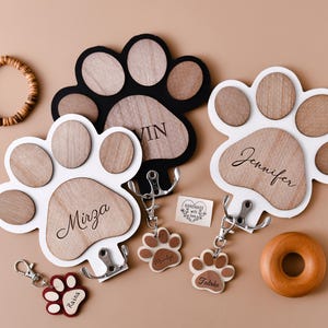 Pode incluir: Três ganchos de chave de madeira em forma de pata de cachorro, brancos e pretos, com os nomes "Mirza", "Jennifer" e "Vin" gravados neles. Cada gancho tem um pequeno chaveiro de madeira em forma de pata de cachorro com os nomes "Raina", "Talulah" e "Murphy" gravados neles.