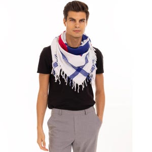 May include: A white square scarf featuring a bold blue and red plaid pattern with a fringed edge. The scarf is draped around the neck, showcasing a prominent blue and red star design on the fabric, with visible white tassels along the border.