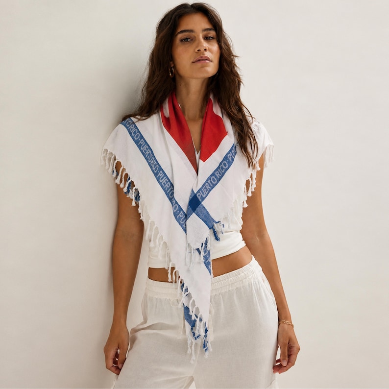 May include: A white triangular scarf featuring a red section at the top and blue stripes along the edges. The blue stripes contain the repeated text PUERTO RICO in a sans-serif font. The scarf has a fringe trim along the bottom edges.