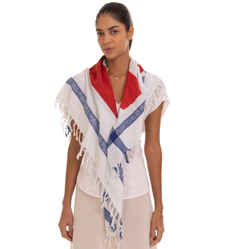 May include: A white square scarf featuring a red triangular accent and blue stripes with the text PUERTO RICO printed repeatedly along the borders. The scarf is finished with white fringe along the edges, creating a textured appearance.