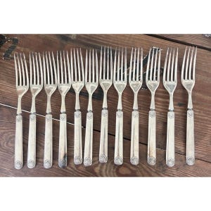 May include: A set of antique silver forks displayed on a wooden surface. Each fork has a decorative handle with a shell design at the base. The tines are slender and evenly spaced, reflecting a classic silverware design.