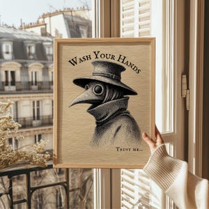 A whimsical plague doctor artwork is elegantly framed in light wood, set against a cozy room with soft natural lighting. Perfect for bathroom wall decor, this piece adds a hint of spooky charm to any space.