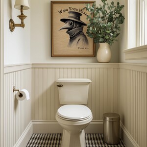 A gold-framed plague doctor art piece with "Wash Your Hands" adds a touch of whimsy to this elegantly styled bathroom. Soft lighting highlights the classic decor, creating a perfect setting for moody bathroom wall art.