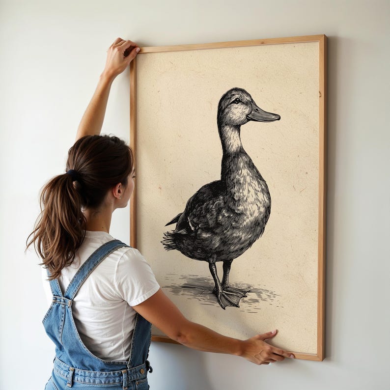 A woman places a vintage duck print in a sleek wooden frame against a light wall, evoking a whimsical farmhouse decor vibe in the softly lit room.