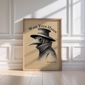 A stylish wooden frame elegantly showcases the spooky plague doctor art with "Wash Your Hands" in bold, positioned against a clean, white-paneled wall, perfect for moody bathroom decor. 

Sunlight filters through, casting intriguing shadows on this whimsical and dark academia-inspired bathroom wall art.