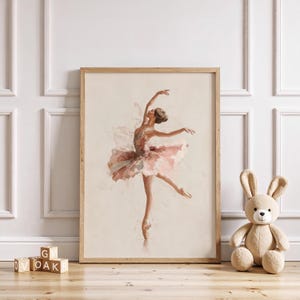 A whimsical ballerina art print in a natural wood frame adds elegance to a girl's room, accompanied by wooden blocks and a plush bunny in a pink nursery setting. The graceful watercolor print brings a dreamy, vintage nursery feel to the space.