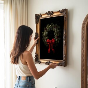 A woman arranges a vintage gold-framed artwork of a festive wreath with a red bow, adding elegance to a cozy, dark academia inspired room adorned with soft curtains and warm lighting.