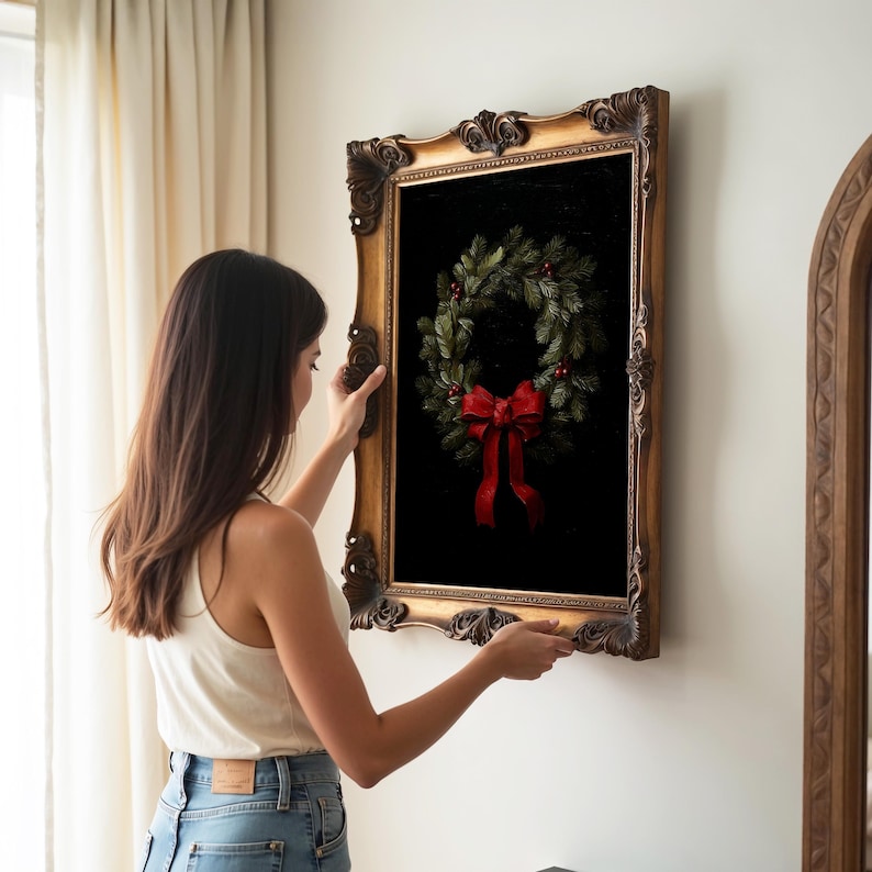 A woman arranges a vintage gold-framed artwork of a festive wreath with a red bow, adding elegance to a cozy, dark academia inspired room adorned with soft curtains and warm lighting.