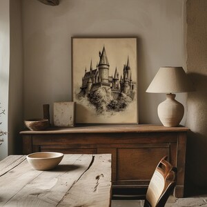 A vintage sketch of a mystical castle hangs in an elegant wooden frame, blending seamlessly with the rustic table and neutral-toned pottery, creating a timeless wizarding decor theme. The cozy lamp light casts a soft glow, enhancing the magical ambiance of this wizarding school art piece.