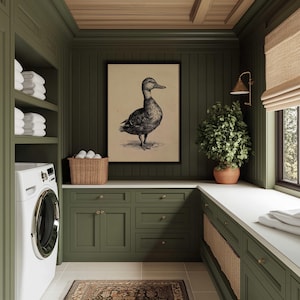 A vintage duck print with a painterly charm is elegantly framed and set against olive cabinetry in a cozy laundry room, accented by wicker baskets and a lush plant, imparting a farmhouse wall decor vibe.