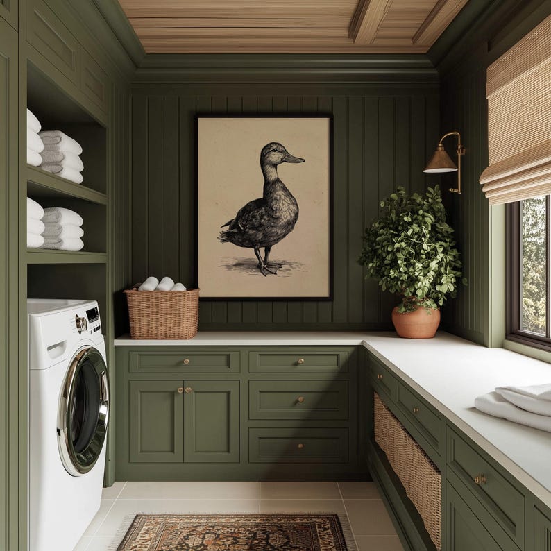 A vintage duck print with a painterly charm is elegantly framed and set against olive cabinetry in a cozy laundry room, accented by wicker baskets and a lush plant, imparting a farmhouse wall decor vibe.