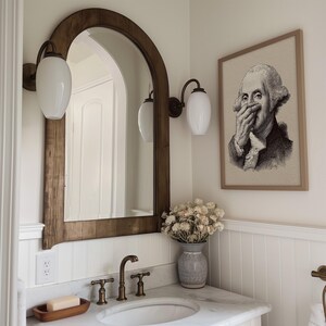 A whimsical vintage sketch of George Washington adds a touch of historical decor to the bathroom, framed elegantly above a marble-topped vanity. Soft lighting highlights the funny bathroom art, complemented by rustic wall sconces and muted floral accents.