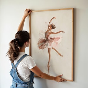 A woman hangs a whimsical watercolor print of a ballet dancer in a natural wood frame, perfect for a ballerina nursery. The delicate ballerina art print adds a touch of elegance to a pink wall decor theme.