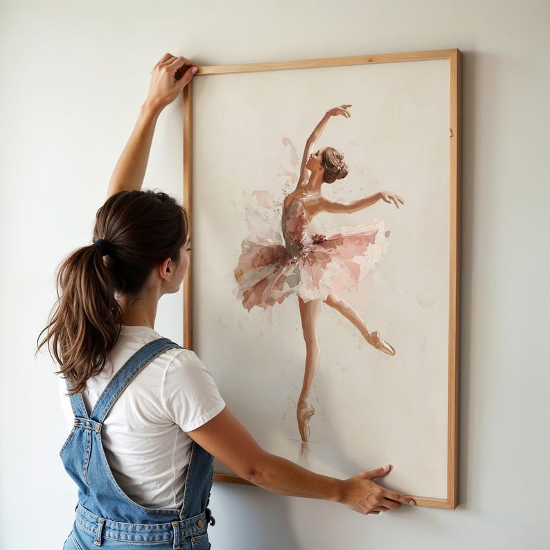A woman hangs a whimsical watercolor print of a ballet dancer in a natural wood frame, perfect for a ballerina nursery. The delicate ballerina art print adds a touch of elegance to a pink wall decor theme.