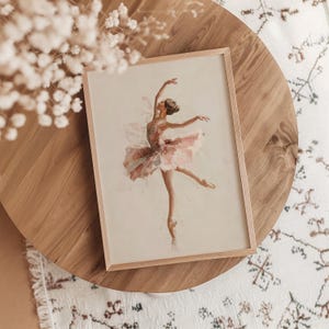 A delicate ballerina art print is framed in light wood, resting gracefully on an oak table, surrounded by subtle floral accents and a patterned rug, perfect for a pink nursery. The watercolor print adds a whimsical touch to a cozy kids room.