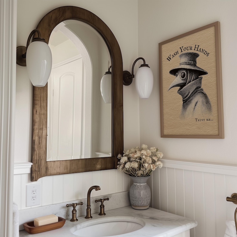 A whimsical plague doctor artwork, titled "Wash Your Hands," adds a touch of funny bathroom art above a marble sink and vintage bronze fixtures. Elegantly framed, it complements the moody bathroom wall decor and muted blooms in a rustic vase.