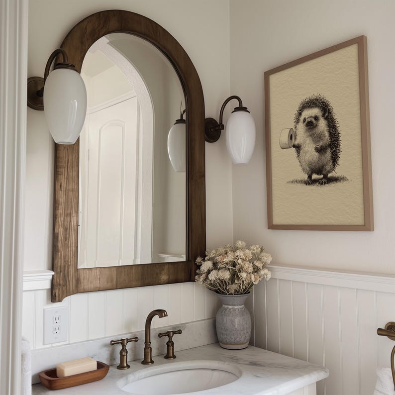 A whimsical hedgehog art print adds a touch of humor to this funny bathroom decor, featuring a rustic wood-framed mirror, elegant brass fixtures, warm lighting, and dried floral accents.