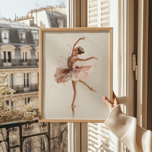 This elegant ballerina art print, framed in light wood, captures a whimsical watercolor ballerina, perfectly complementing the soft light of a vintage nursery. Displayed against a Parisian window, the charming pink wall decor brings sophistication to a ballerina nursery.