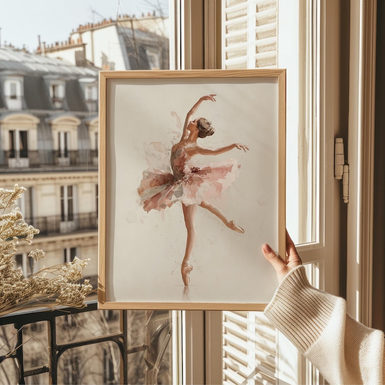 This elegant ballerina art print, framed in light wood, captures a whimsical watercolor ballerina, perfectly complementing the soft light of a vintage nursery. Displayed against a Parisian window, the charming pink wall decor brings sophistication to a ballerina nursery.