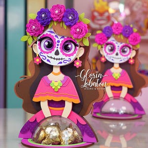 May include: Two decorative paper dolls in the style of Día de Muertos. Each doll features a sugar skull face, a floral crown, and a pink dress. The dolls are holding a clear dome with gold-wrapped candies. The text "Gloria Lobaton" is visible.
