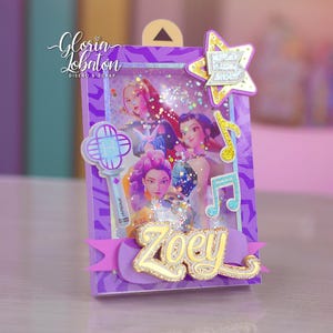 May include: A purple shadow box with a picture of a group of women. The box has a star, musical notes, and the name "Zoey" in gold. The box is decorated with glitter and purple ribbon.