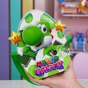 Puede incluir: Decoración de fiesta con temática de Yoshi, verde y blanca, con el personaje saliendo de un huevo decorado. La decoración incluye dos estrellas amarillas y las palabras "Mario Galaxy" en una fuente colorida.